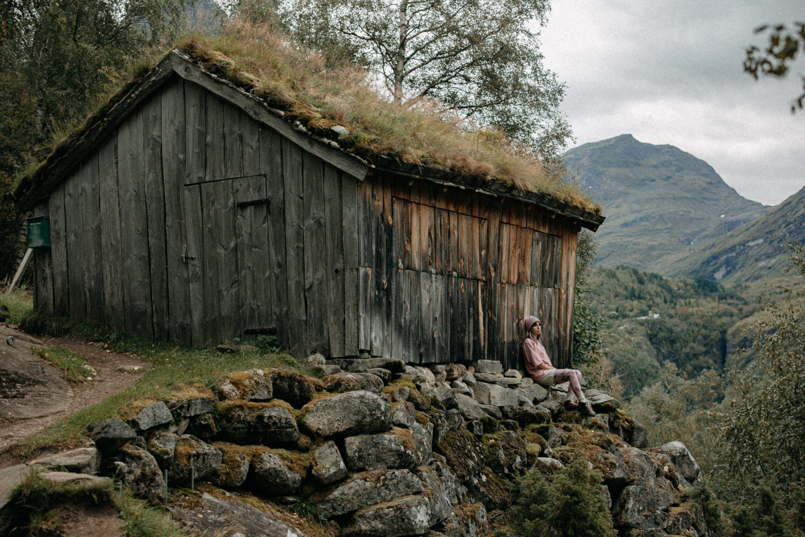 Woman in soft pink clothes sitting by a weathered wooden cabin, nestled in a mossy mountain landscape — a quiet, dreamy pause in nature.