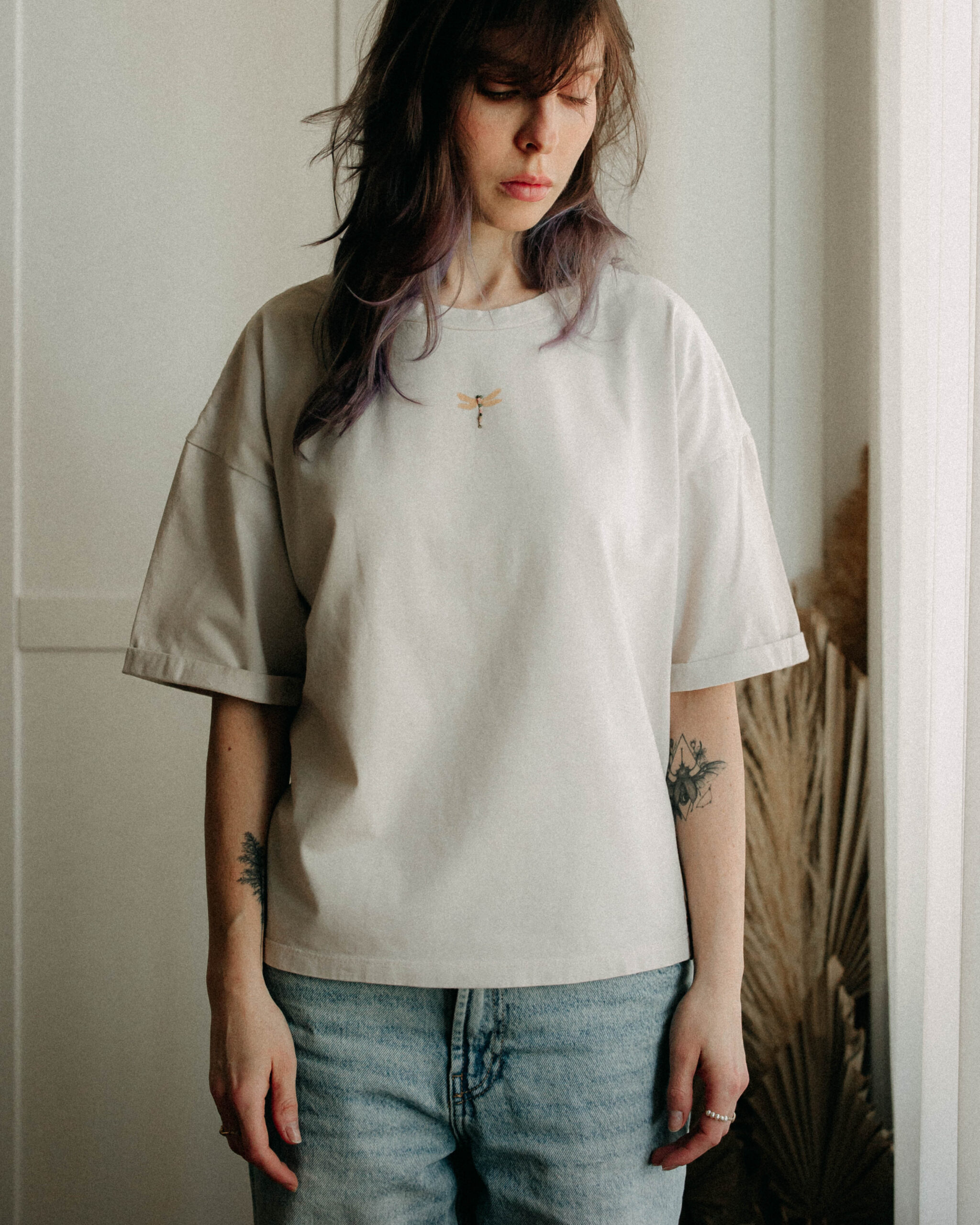 Woman standing in soft daylight wearing an oversized pale tee with a small dragonfly print.