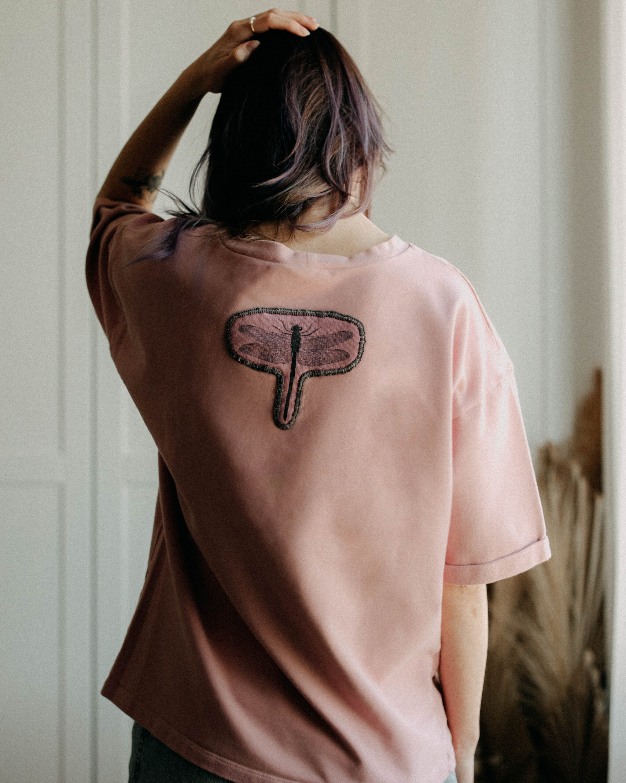 Back view of a woman wearing a blush pink tee with a small dragonfly print.