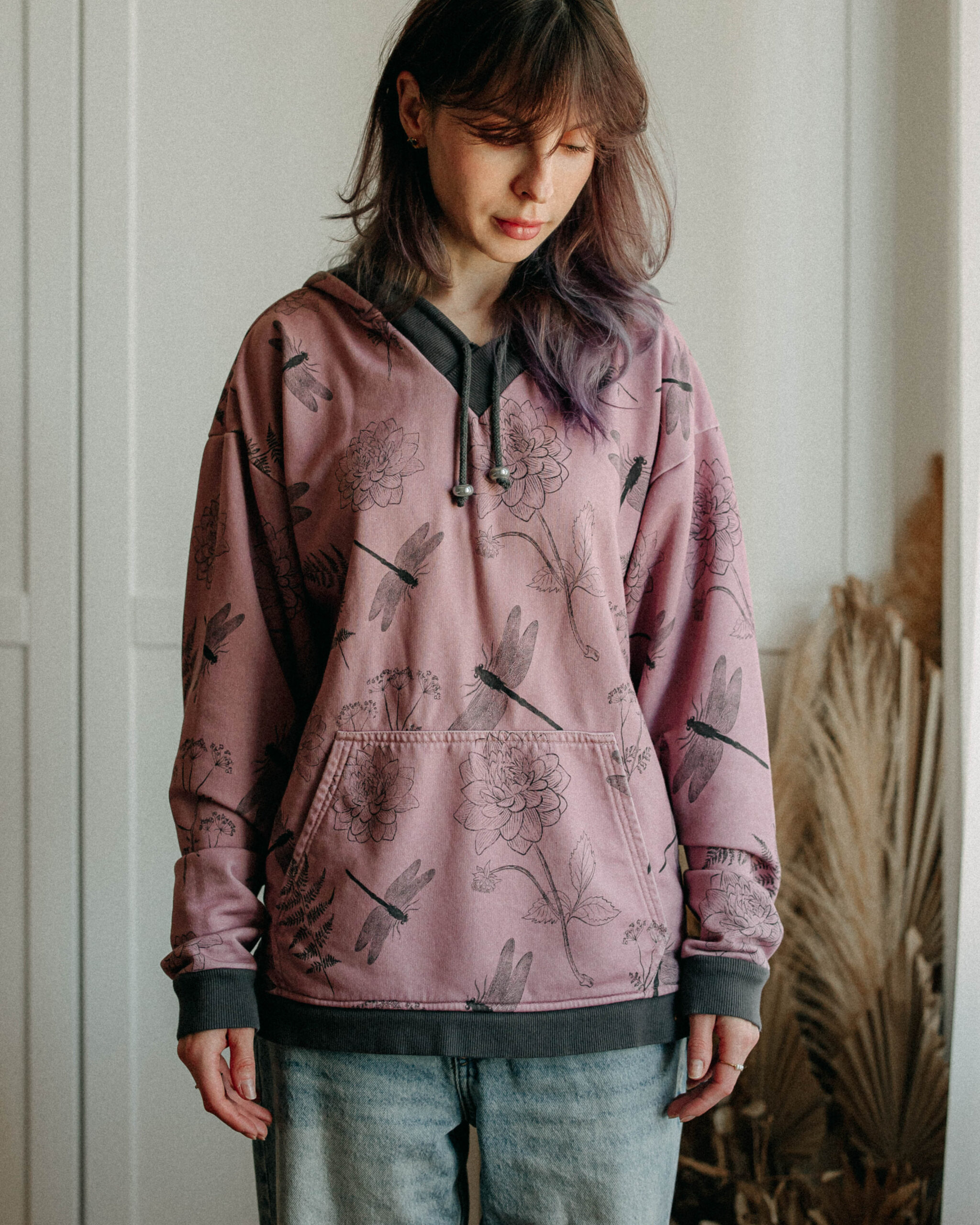 Woman wearing a pink hoodie with floral and dragonfly illustrations, standing indoors.