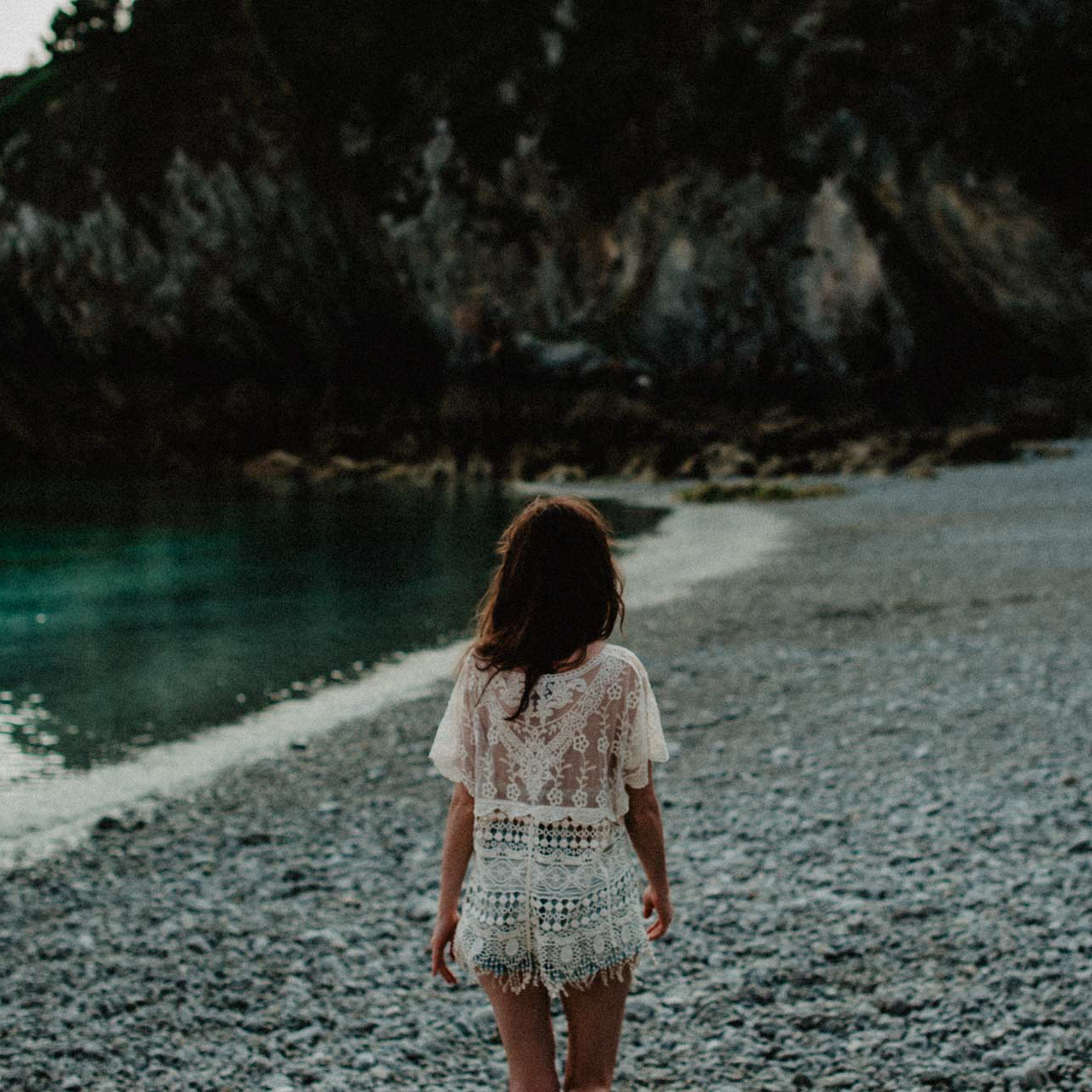 Woman walking barefoot on a rocky beach, facing the turquoise water and distant cliffs, wearing a lace dress under soft afternoon light.