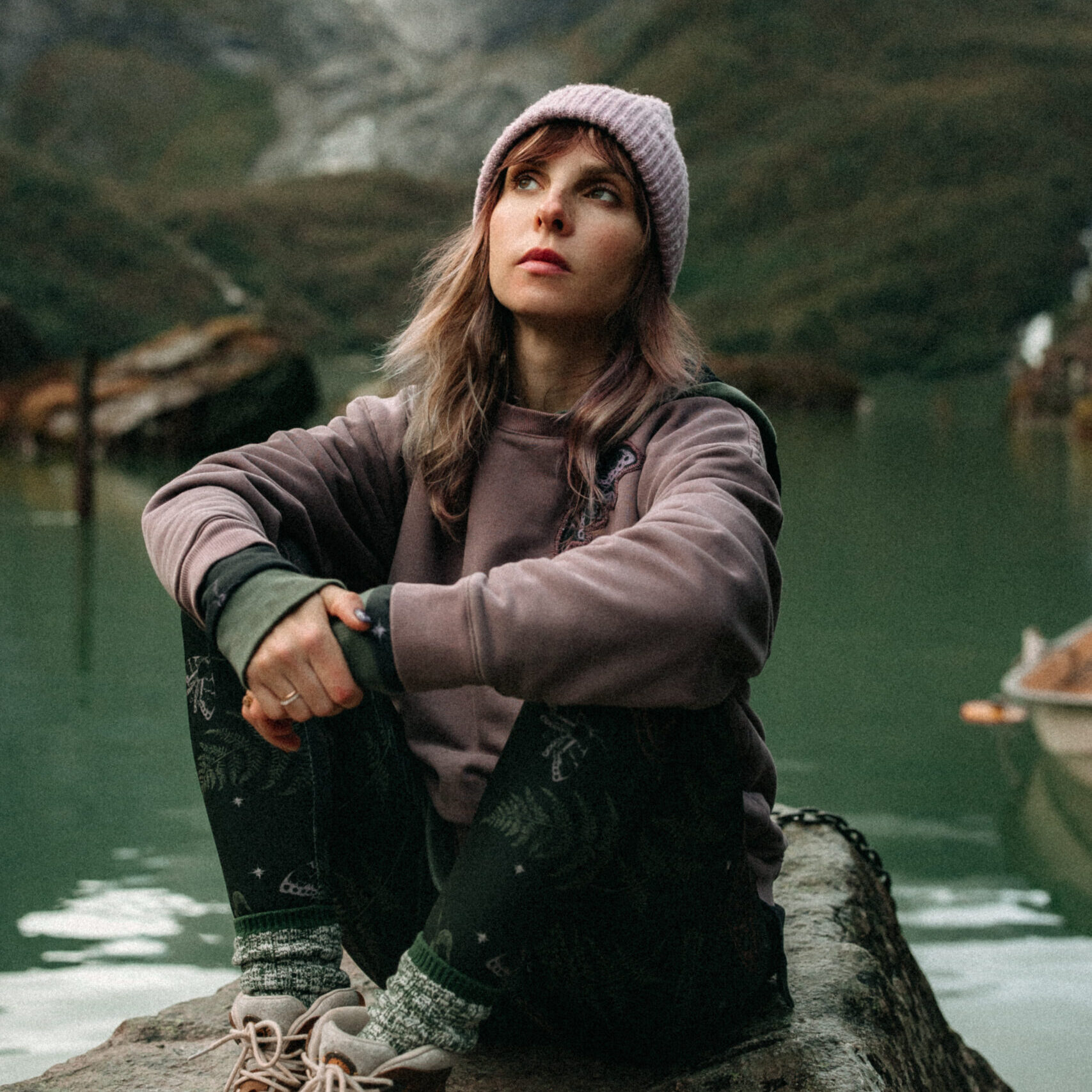 Woman in a forest green hoodie and pink beanie sits cross-legged on a lakeside rock, looking upward with serene determination.