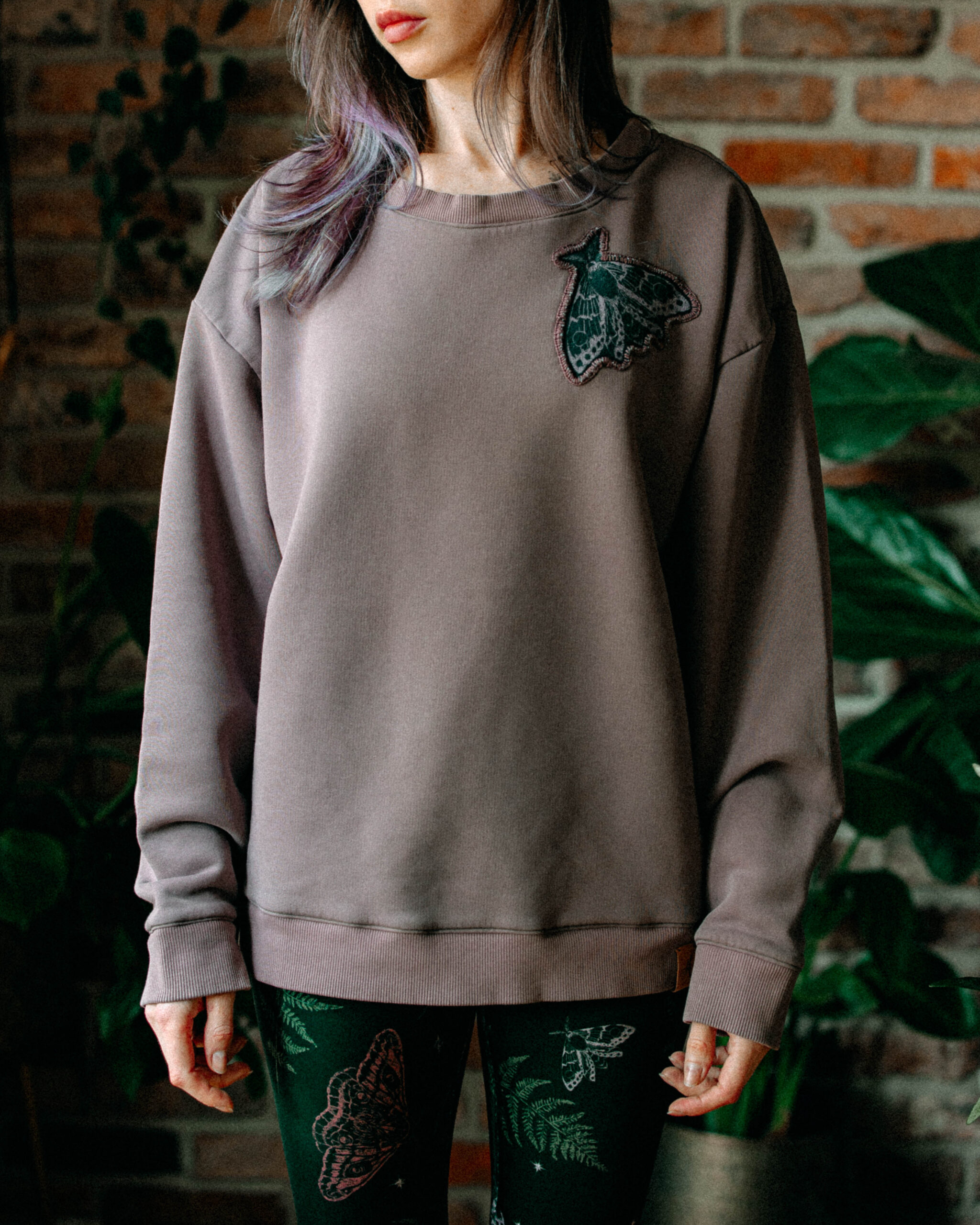 Woman wearing a mauve-gray sweatshirt with a green moth appliqué, standing against a brick wall.