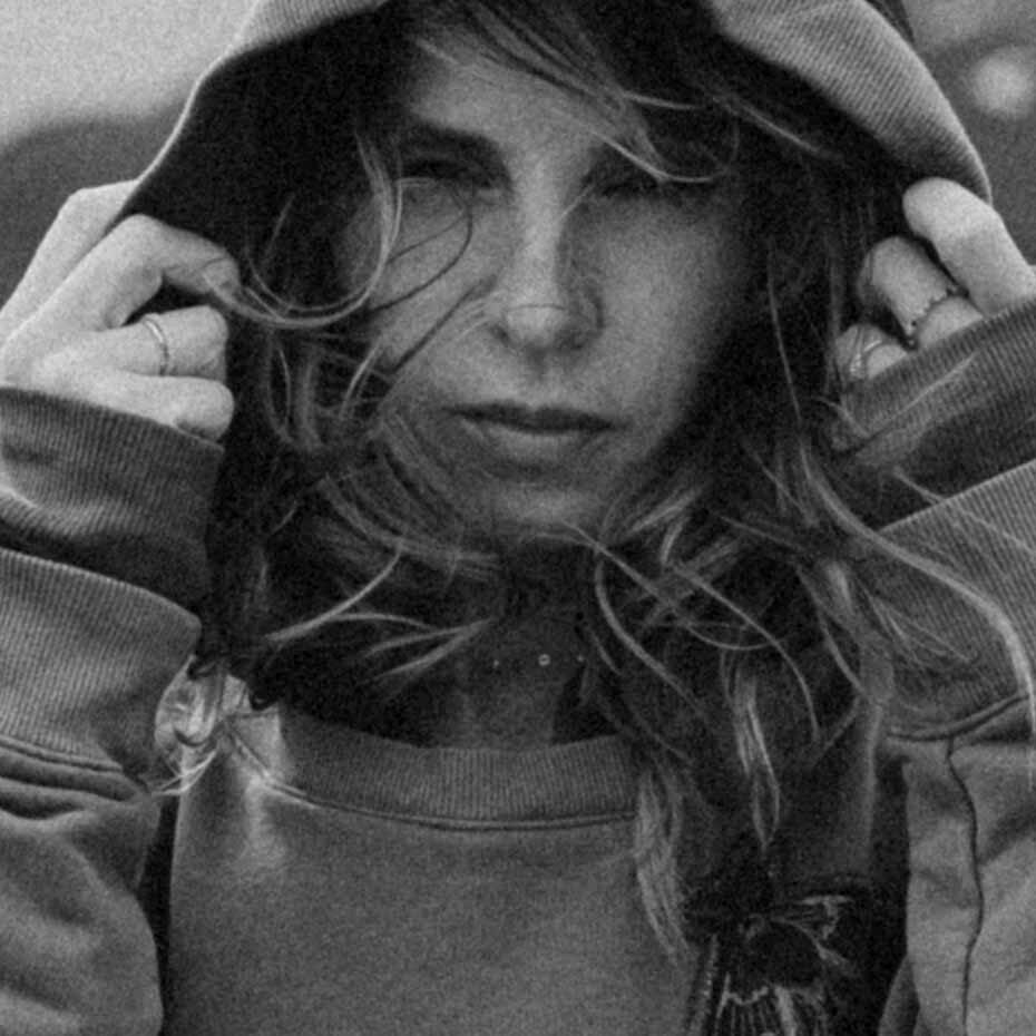 Black and white portrait of the Ferny Tales founder wearing a hoodie with the hood up, holding the edges softly, gazing forward with windblown hair.