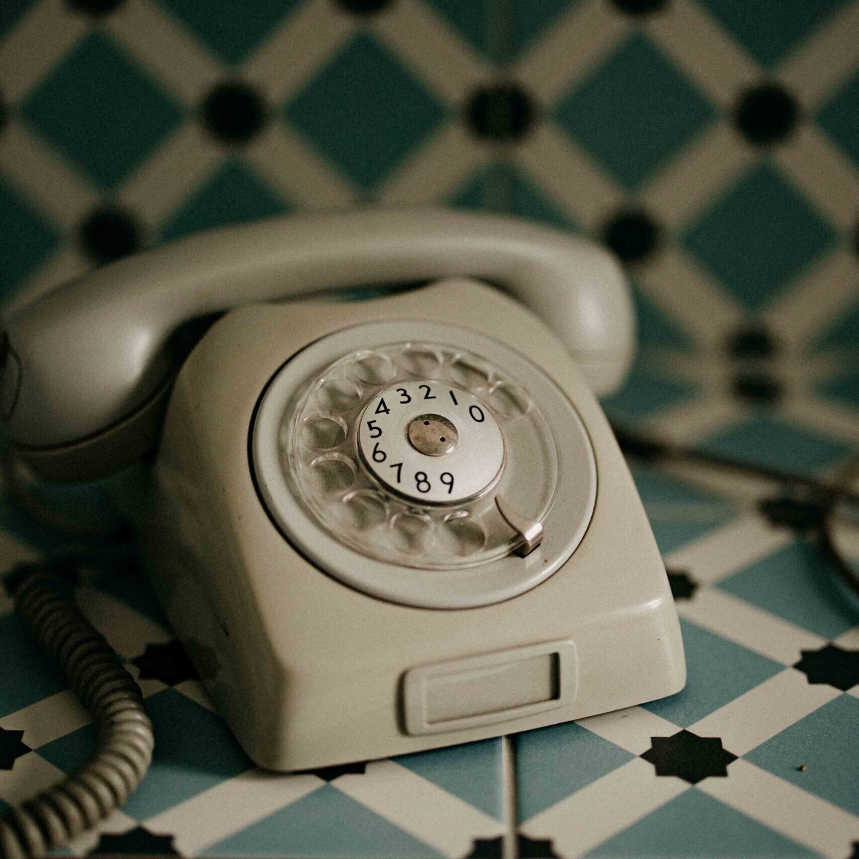 A pale rotary dial phone resting on a patterned teal-and-white tile surface, evoking a sense of nostalgic calm.
