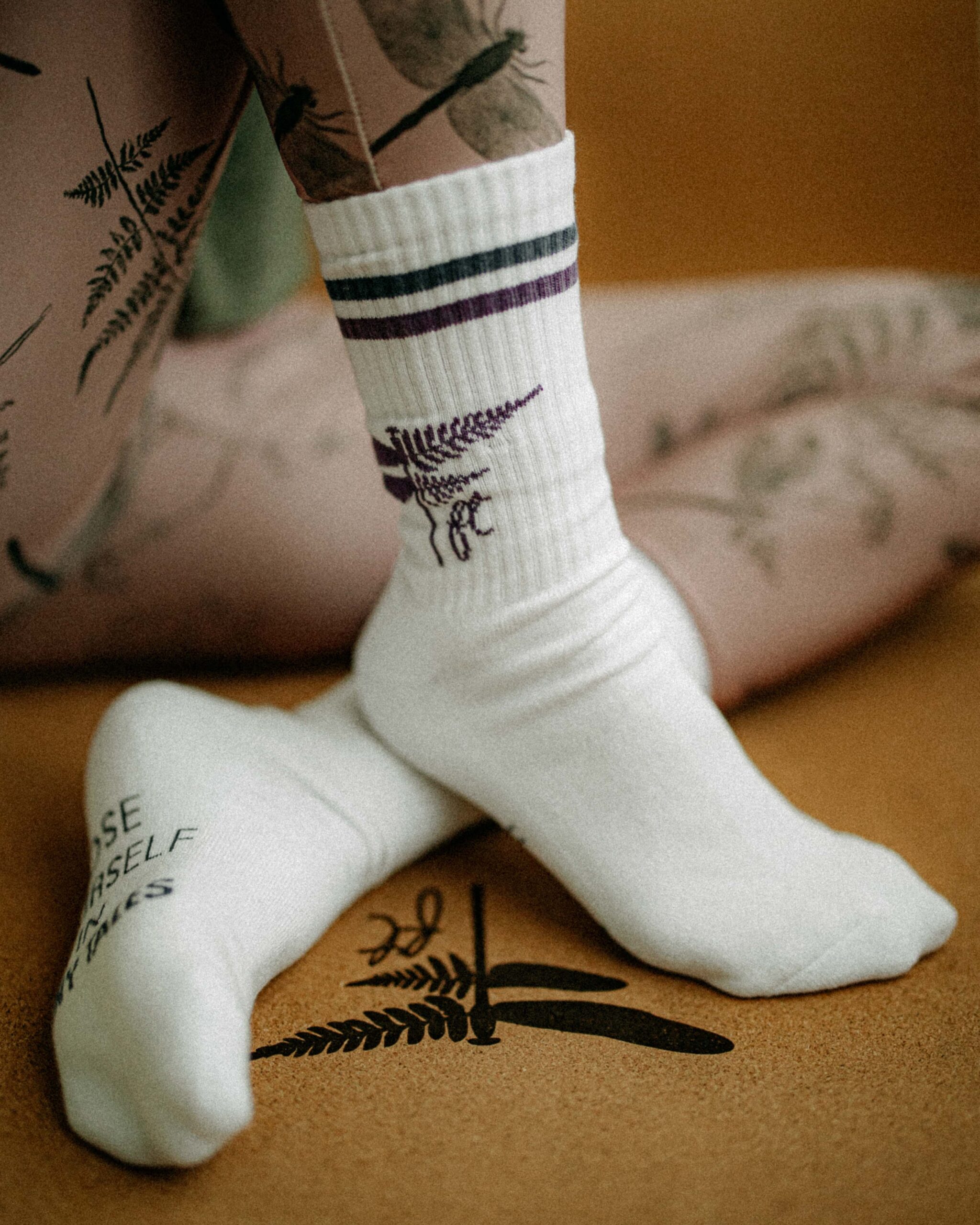A close-up of Ferny Socks worn over fern-printed leggings. The white ribbed fabric features a woven fern and dragonfly motif in aubergine and navy, captured against a soft indoor backdrop with botanical tattoos visible.