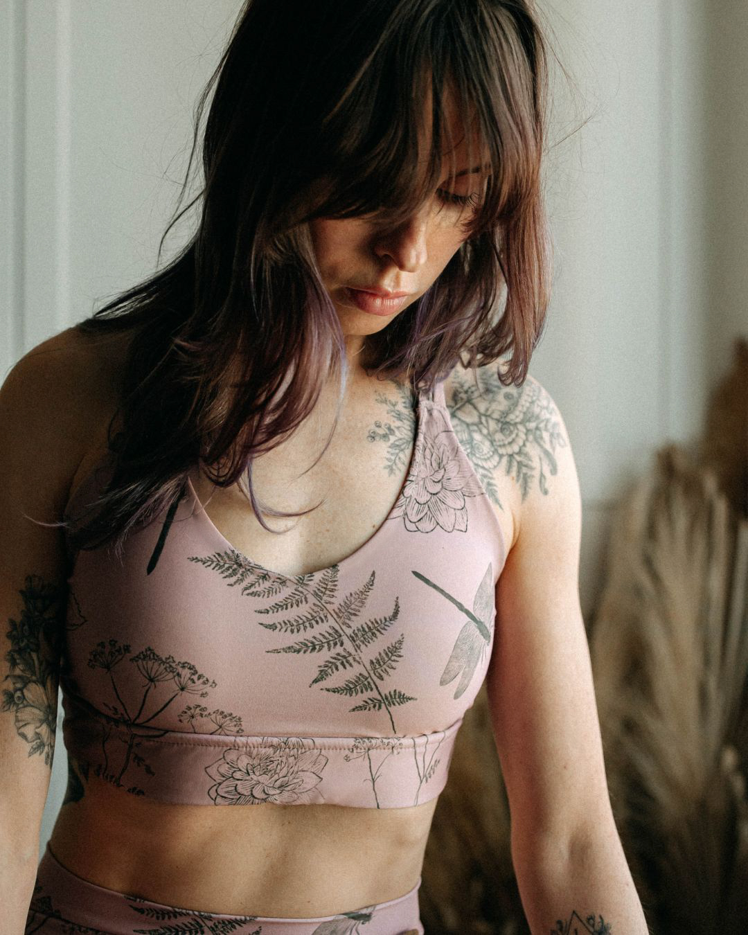 Woman in a pale pink sport top with garden illustrations, standing by a window.